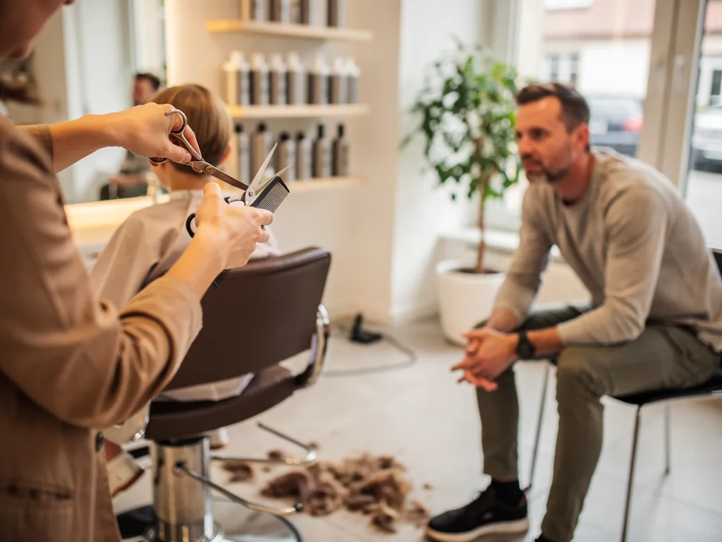 Friseurin schneidet einem Kind im Salon die Haare, während der Vater daneben sitzt und zusieht.