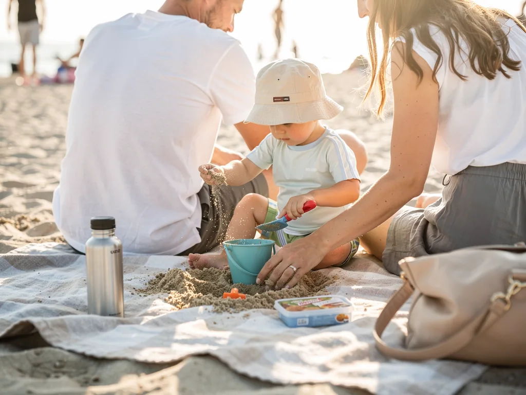 Kleinkind mit Sonnenhut spielt zwischen seinen Eltern auf einer Stranddecke mit Sand, Eimer und Schaufel.