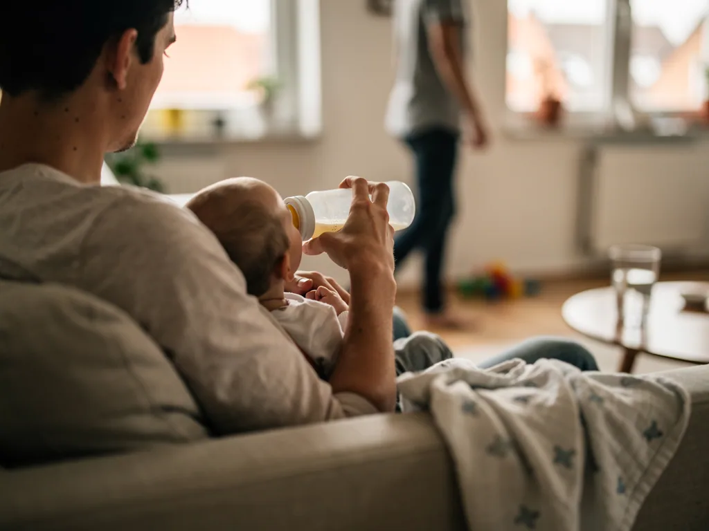 Vater füttert ein Baby auf dem Sofa mit der Flasche, im Hintergrund steht eine zweite Person am Fenster.