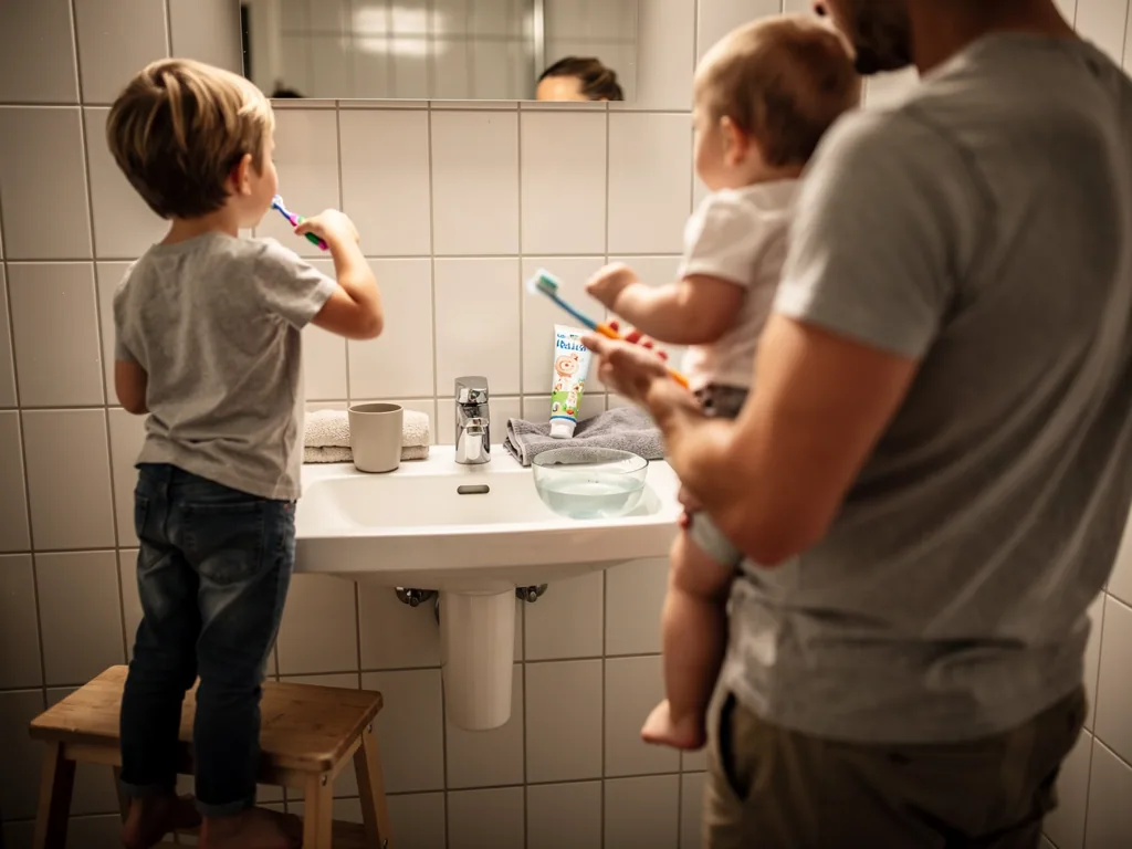 Vater hält Baby mit Zahnbürste, während ein älteres Kind am Waschbecken die Zähne putzt.