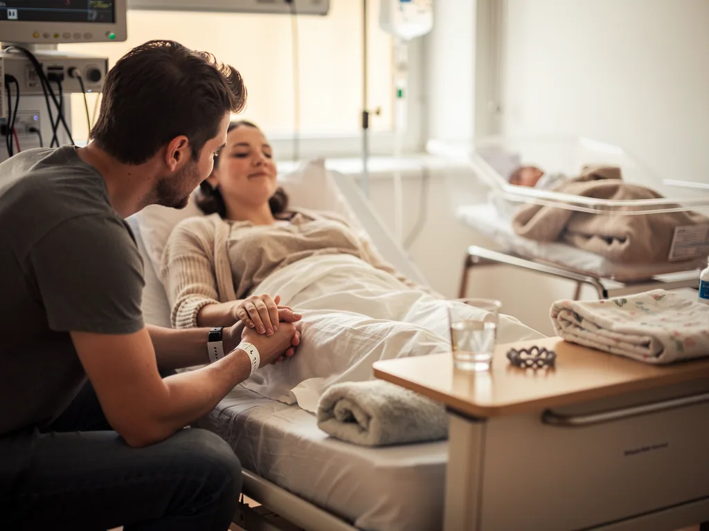 Vater hält die Hand der Mutter im Klinikbett; daneben liegt ein Neugeborenes im Babybettchen.