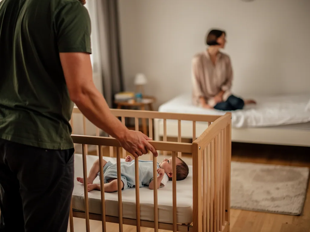 Vater steht am Babybett mit wach liegendem Säugling, Mutter sitzt im Hintergrund auf dem Bett.