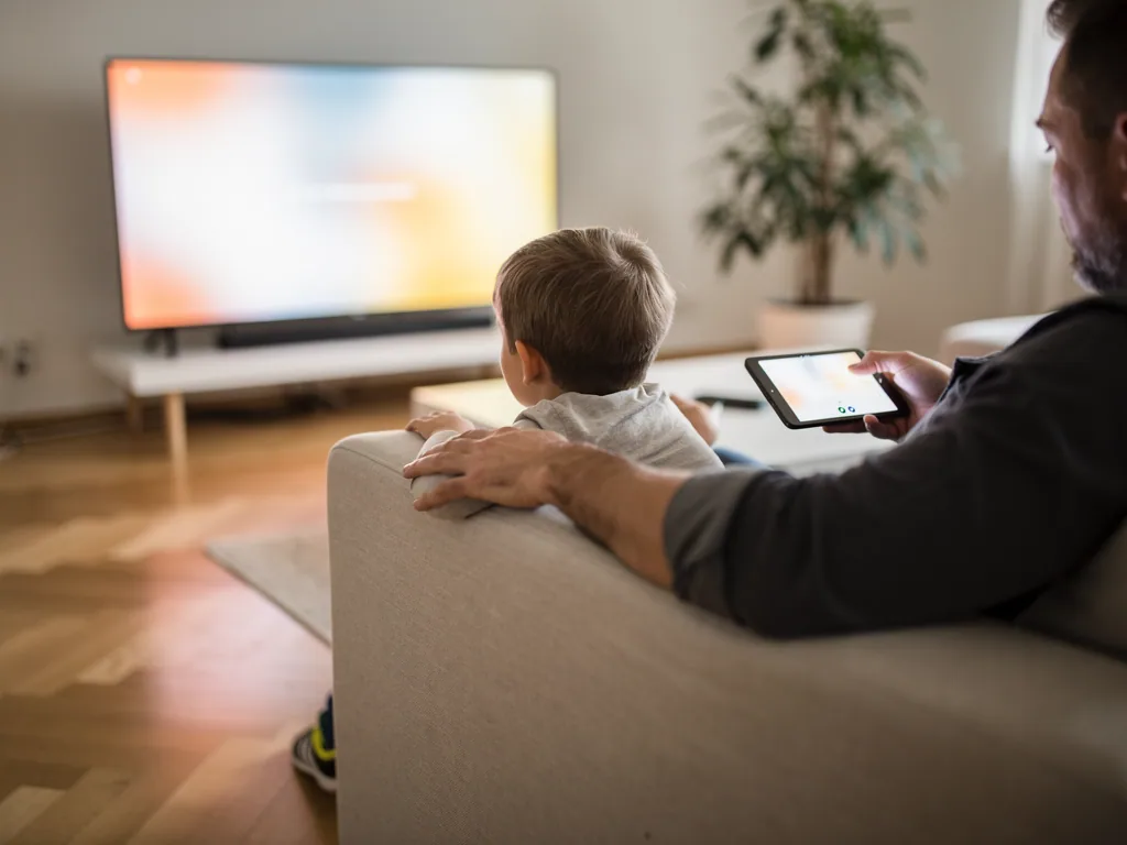 Vater und Kind sitzen auf dem Sofa vor dem Fernseher; der Vater hält ein Tablet in der Hand.